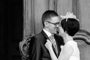 Hochzeitspaar lehnt an einer alten großen Holztür, schwarzweiß-fotografie, lächelt sich gegenseitig an, die Nasenspitzen berühren sich