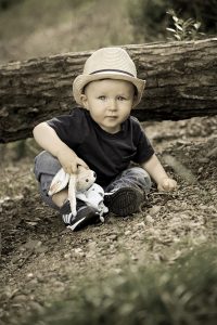 Kleinkind mit Teddy und Hut sitzt auf Waldboden