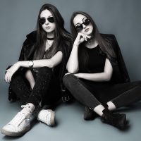 Zwei junge Frauen, fast komplett schwarz gekleidet sitzen mit dunklen Sonnenbrillen leger auf dem Studioboden