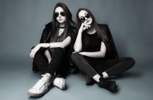 Zwei junge Frauen, fast komplett schwarz gekleidet sitzen mit dunklen Sonnenbrillen leger auf dem Studioboden