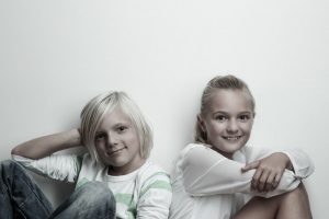 Blondes Geschwisterpaar: Junge und Maedchen sitzten Rücken an Rücken, blicken direkt in die Kamera, lächeln
