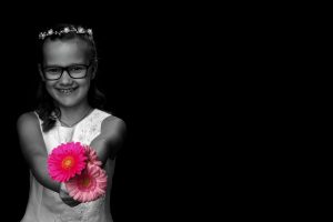 Junges Mädchen lächelt direkt in die Kamera und streckt 3 Gerbera in die Kamera, die Blumen sind in leuchtenden rot-rosa Farben dargestellt, das Maedchen ist in schwarz-weiß dargestell,t, schwarzer Hintergrund
