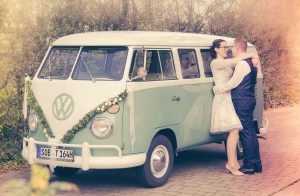 Hochzeitspaar steh an einem alten VW-Bus gelehnt, sich anblickend und sich umarmend