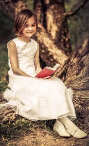 Portraitfoto eines jungen Mädchen in weißem Kleid, mit Gebetbuch in der Hand, in die Kamera lächelnd